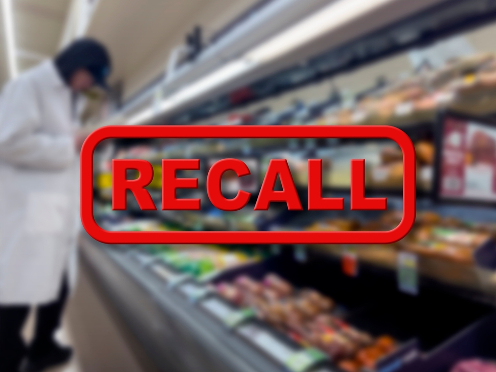 Blurred background of a grocery store employee removing recalled product with the red text Recall in the foreground
