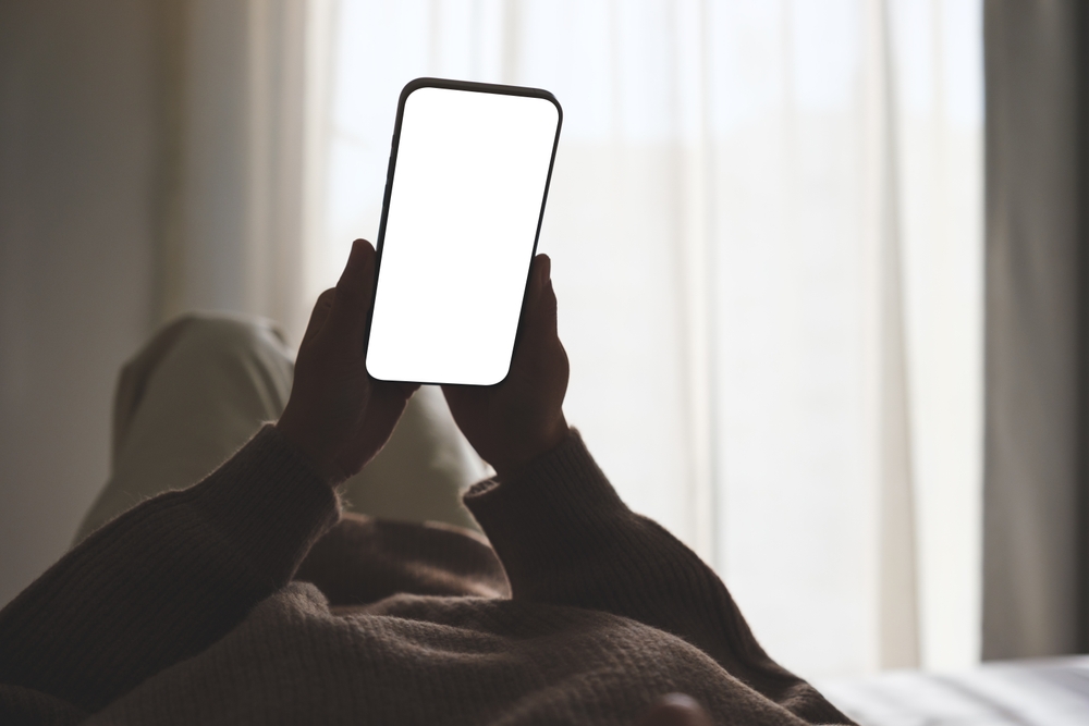 Mockup image of woman holding mobile phone with blank desktop white screen while lying on bed