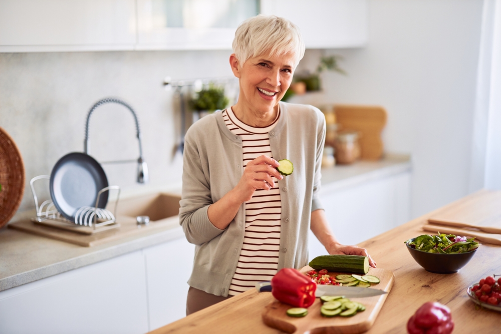 A portrait of a lovely and happy senior adult woman with short hair leaning on a kitchen counter and eating a slice of cucumber while preparing a healthy salad.