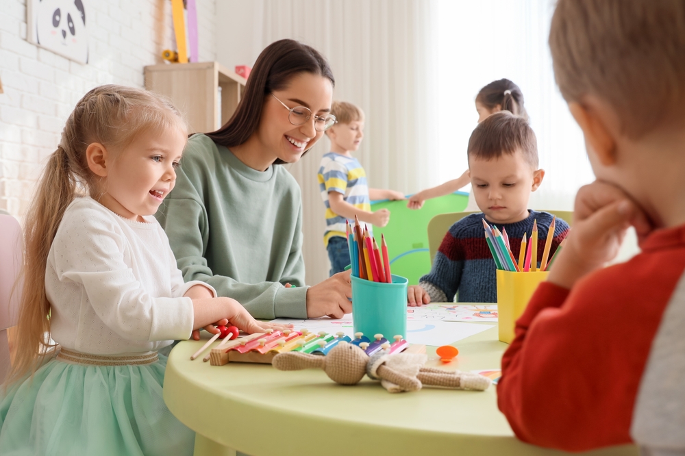 Cute little children with nursery teacher drawing at table in kindergarten
