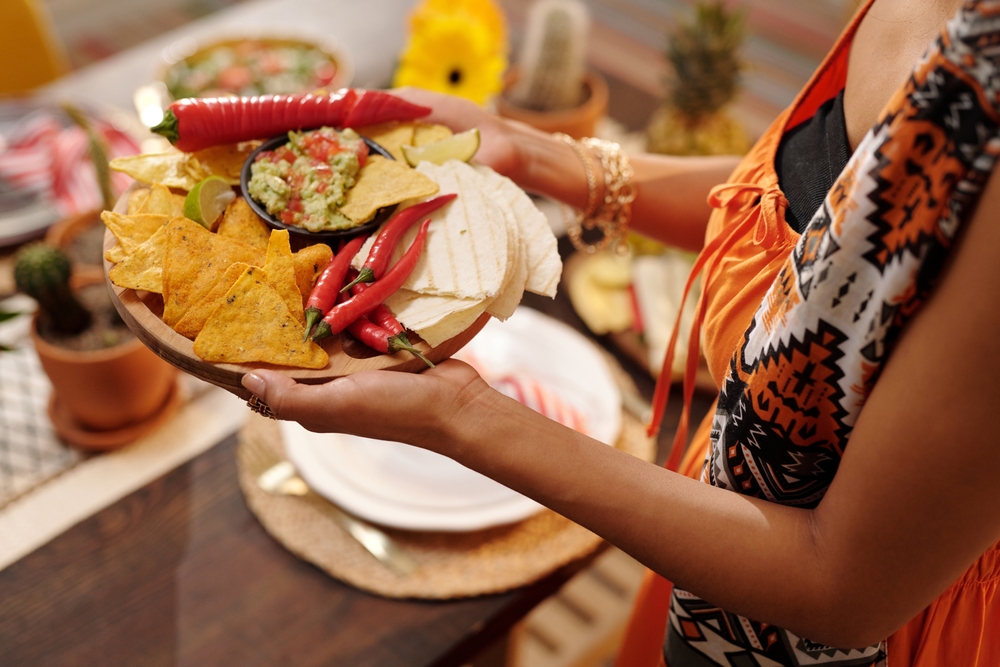 Tray with spicy chips, sauce, tacos and red hot chili peppers held by young Hispanic woman in national attire serving table for festive dinner