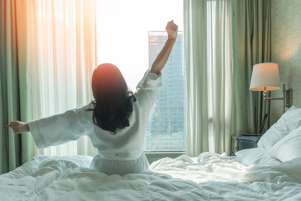 Hotel relaxation on lazy day with Asian woman waking up from good sleep on bed in weekend morning resting in comfort bedroom looking toward city view, having happy, work-life quality balance lifestyle

