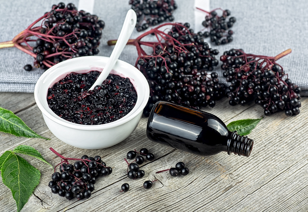 Delicious homemade black elderberry syrup in glass jar and bunches of black elderberry with green leaves on wooden desk. Sambucus.
