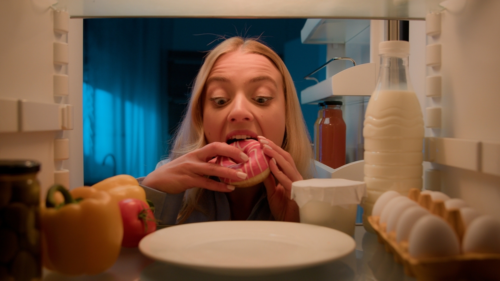 Point of view POV from inside refrigerator hungry Caucasian woman girl at night evening kitchen open fridge take out eating sweet donut unhealthy nutrition obesity diet failure overeating food addict