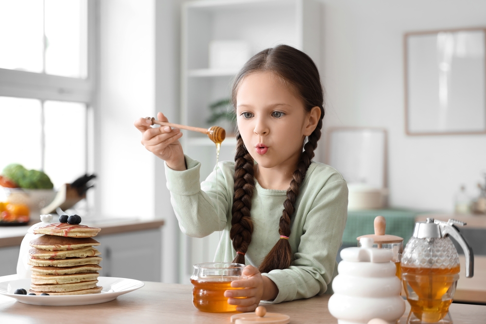 Cute little girl with honey and pancakes at table in kitchen