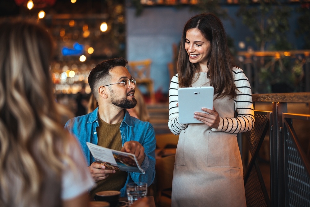 A happy female waitress wearing an apron interacts with dining guests, taking orders on a digital tablet in a cozy restaurant setting.
