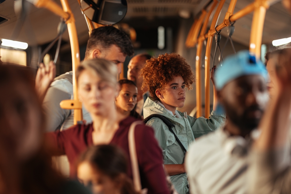 Teenage girl with curly hair standing on a crowded public bus among other passengers.
