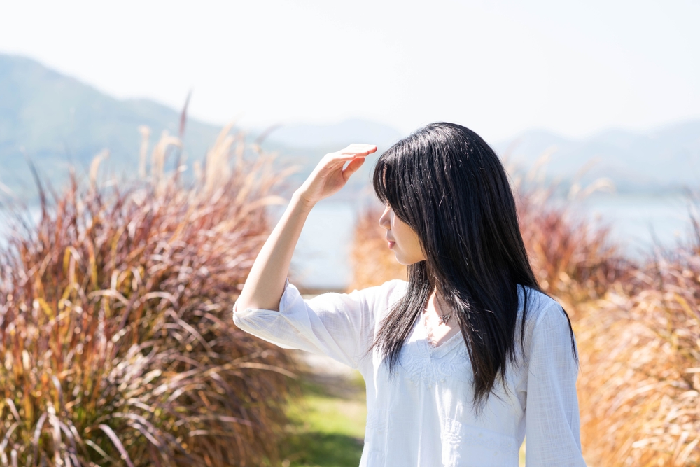 Portrait of a beautiful Asian girl with long black hair covering face by hand of bright sun light. Teenager girl in a white dress protecting her face from solar light. Skin care or beauty concept.
