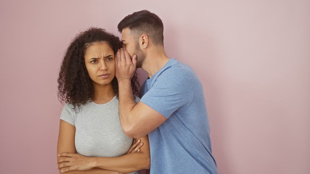 A man whispers into a skeptical woman's ear against a pink background, suggesting secrecy or gossip.
