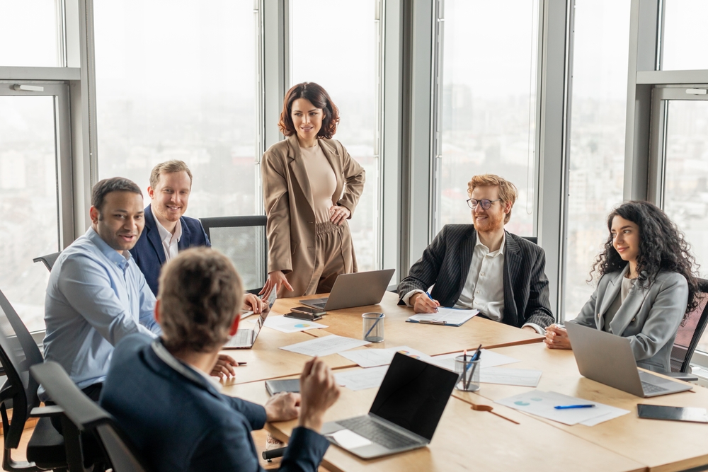 A group of diverse business professionals are seated around a large conference table in a modern office space. One woman stands at the head of the table, while the others focus on the discussion