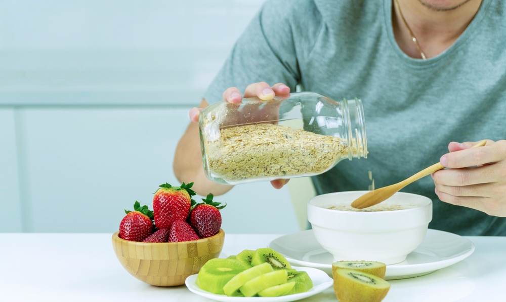 A person is pouring oatmeal into a bowl while holding a jar of oatmeal. The bowl is filled with fruit, including strawberries and kiwi. Concept of health and wellness
