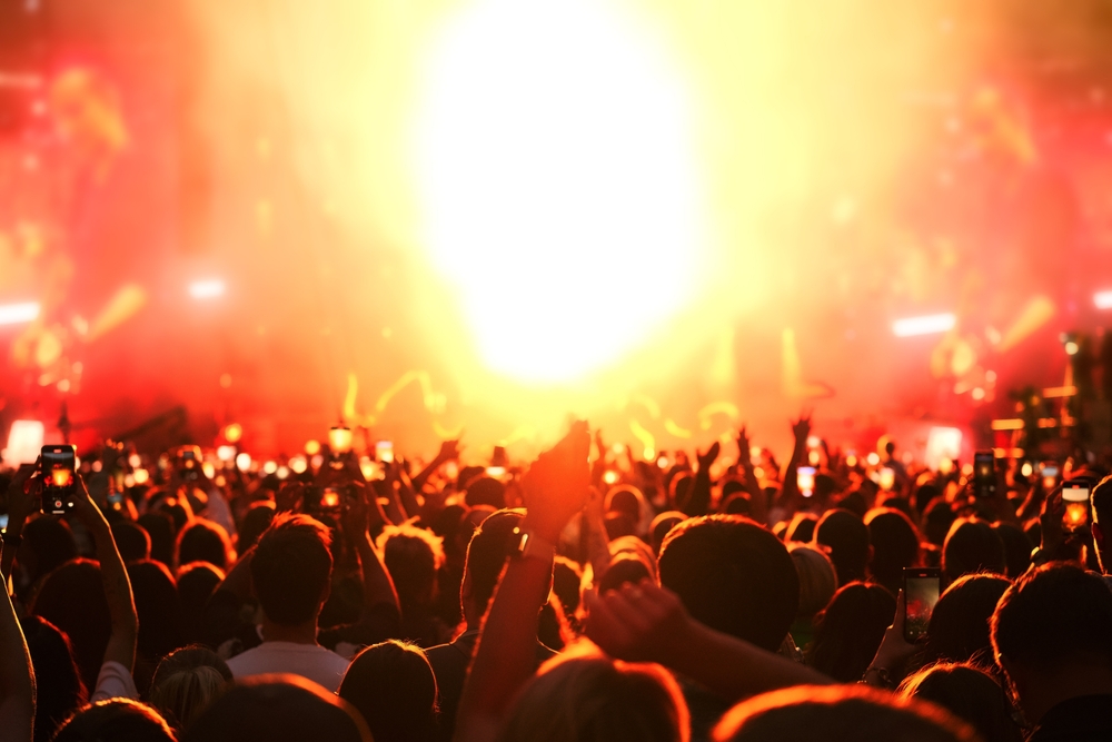 Crowd of people enjoys and cheering music concert in front of bright stage lights. Happy audience dance and raise hands.
