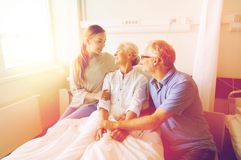 medicine, support, family health care and people concept - happy senior man and young woman visiting and cheering her grandmother lying in bed at hospital ward