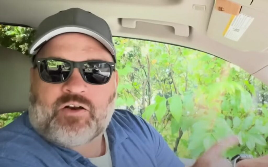 A man sits in the driver seat of his car. Greenery in the background.