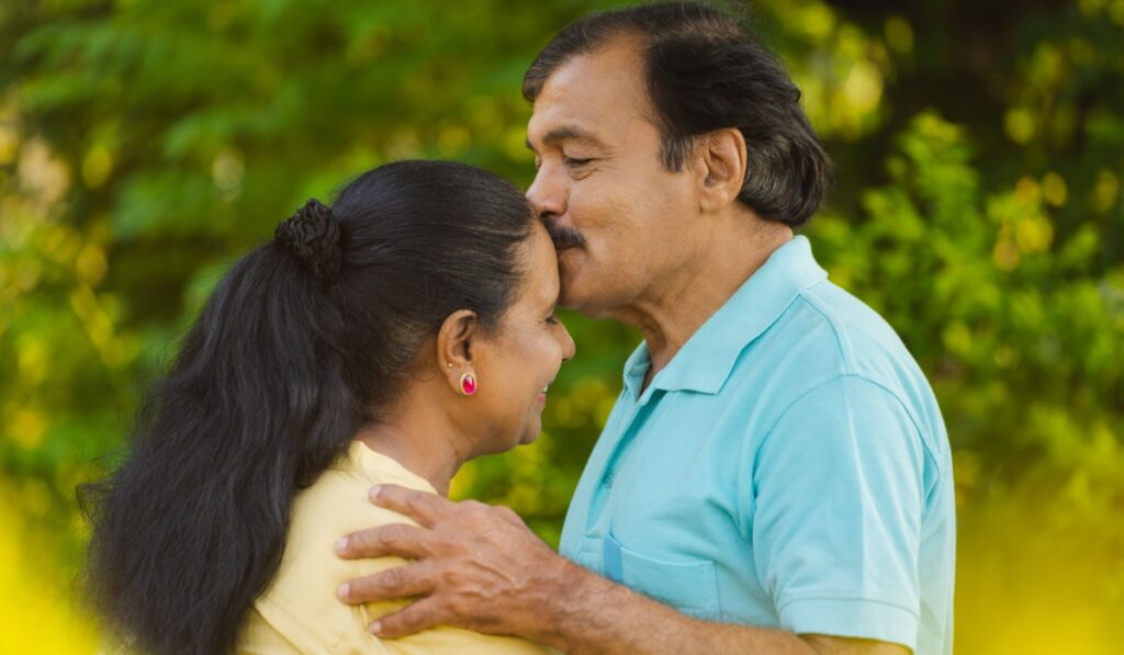 Happy senior man or husband kissing his wife's forehead by hugging at park - concept of emotional bonding, physical intimacy and romantic moment.