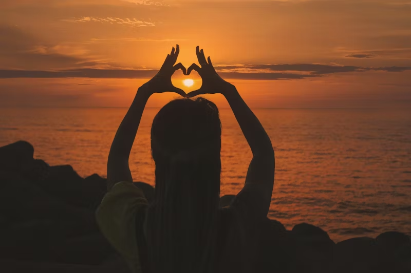 woman making a heart symbol around the setting sun
