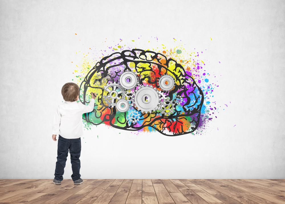 Rear view of a cute little boy wearing a white shirt and dark blue jeans writing or drawing with a marker. A concrete wall background with a cog brain sketch on it.