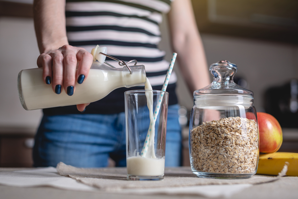 A woman pours organic oat milk from a bottle into a glass on a table in the kitchen. Diet healthy vegetarian product