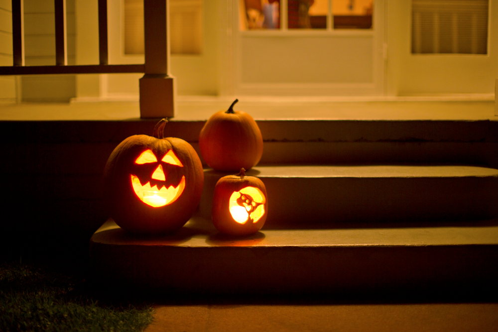 Two jack o' lanterns glowing on the steps of a house beside a normal pumpkin.