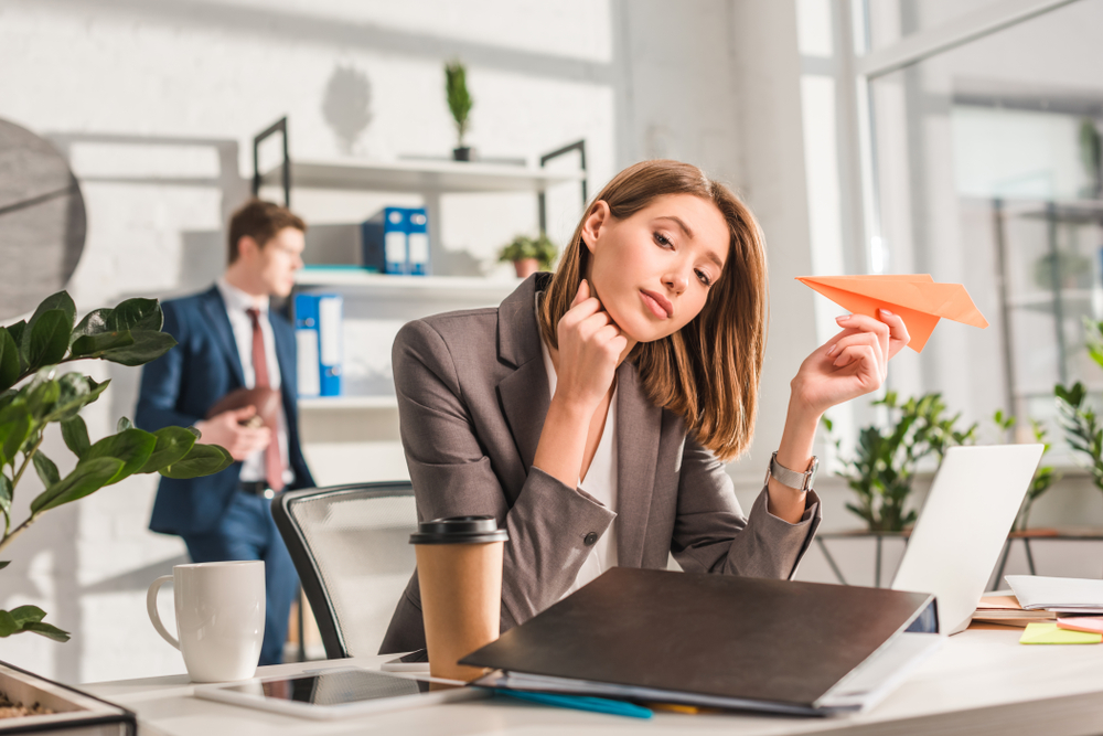 tired businesswoman holding paper plane near laptop with coworker on background, procrastination concept
