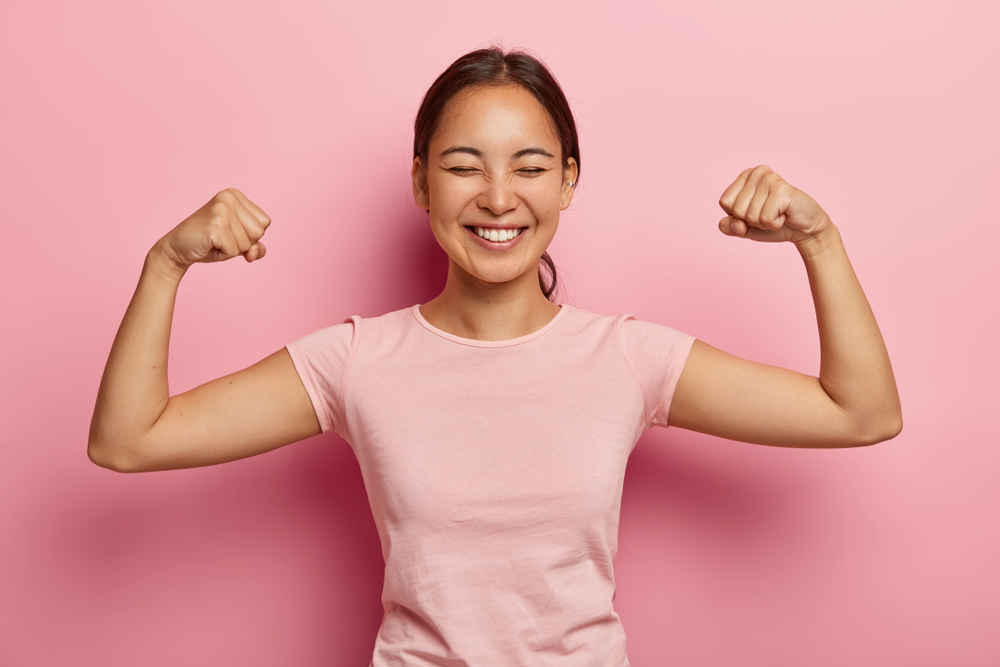 Strong powerful Asian woman with dark combed hair, toothy smile, raises arms and shows biceps, has piercing in ear, wears casual rosy t shirt, models against pink background. Look at my muscles!