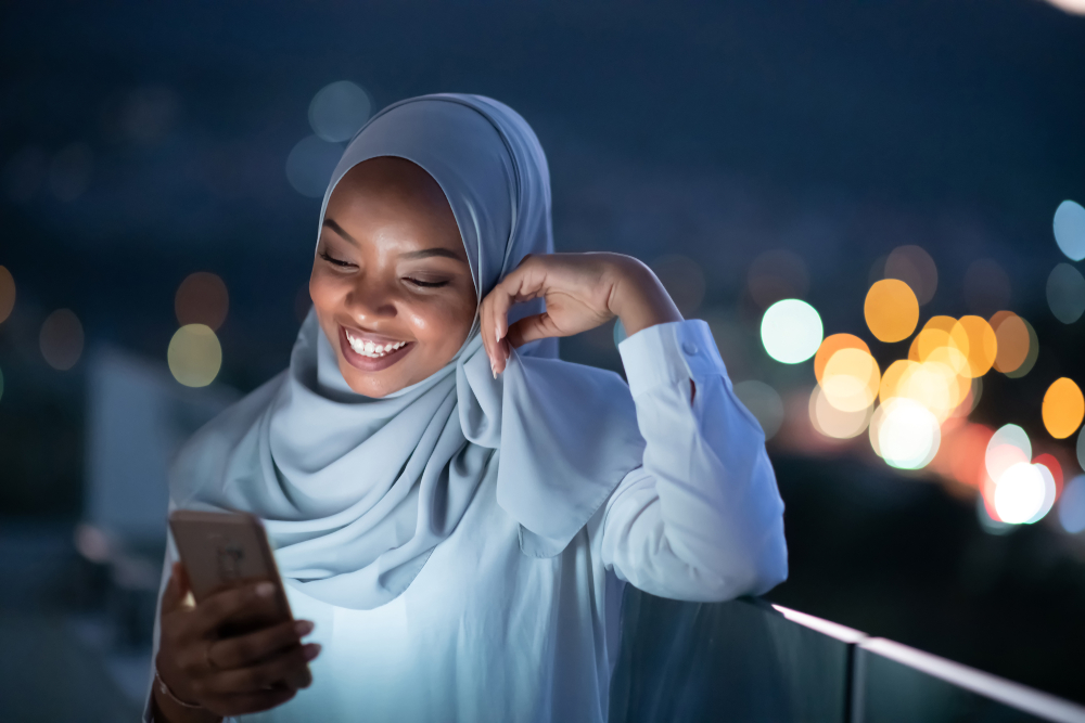 Young Muslim woman wearing scarf veil on urban city street at night texting on smartphone with bokeh city light in background
