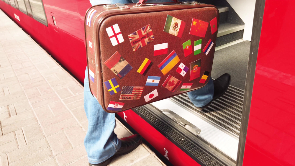A man with a vintage suitcase comes into the vestibule of the train. The suitcase is decorated with stickers with the image of flags of different countries.