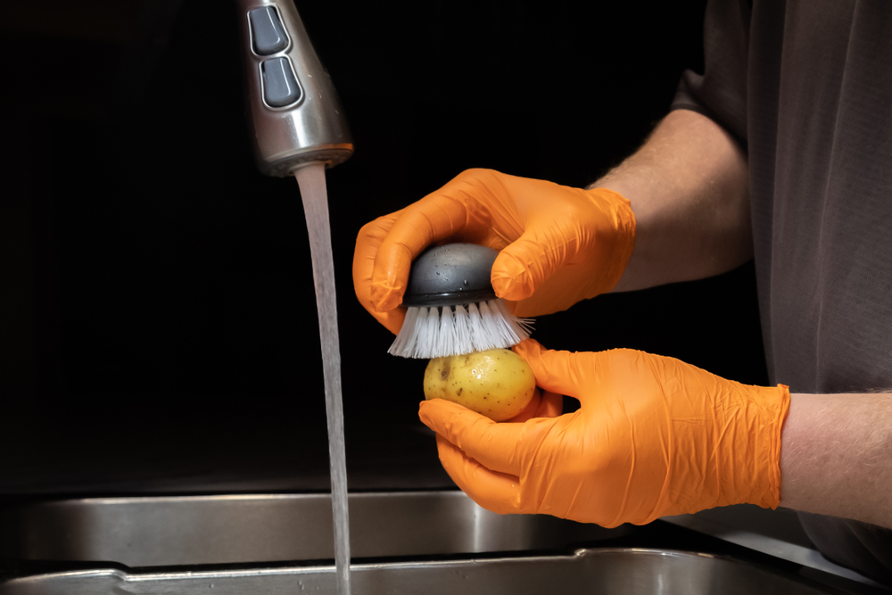 Close up of washing and rinsing potatoes in preparation for cooking. Model wearing orange disposable rubber gloves.

