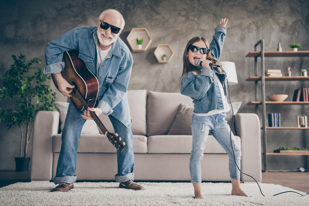 Photo of two people funky grandpa play guitar small nice granddaughter mic singing cool style sun specs denim clothes repetition school concert stay home quarantine living room indoors