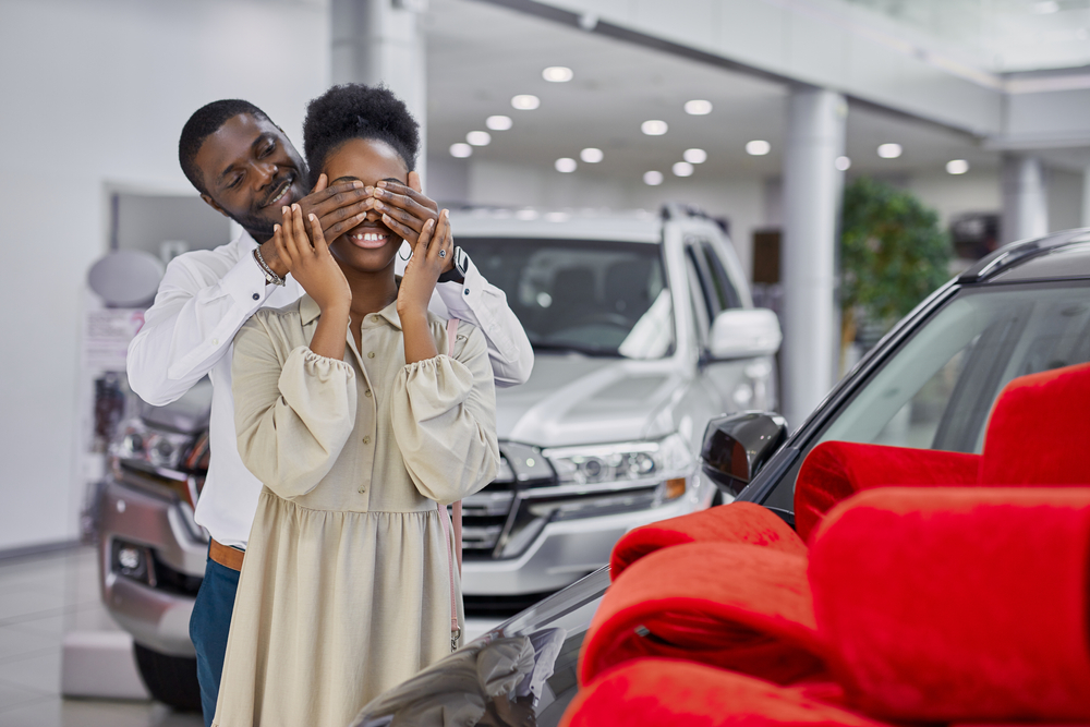 black man prepared gift to his wife, going to give new auto as a present, man closed woman's eyes during surprise. luxurious car is wrapped in red bow, in dealership
