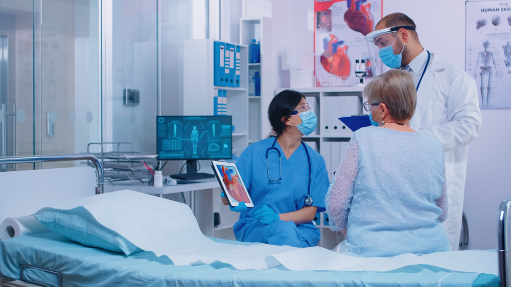 Nurse in mask with cardiac patient at doctor appointment showing a heart representation on digital tablet in modern clinic. Doctor entering hospital room talking with old retired senior woman patient
