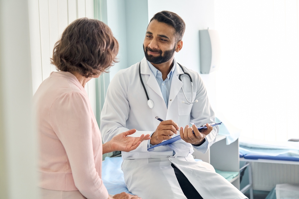 Indian male doctor consulting senior old patient filling form at consultation. Professional physician wearing white coat talking to mature woman signing medical paper at appointment visit in clinic.
