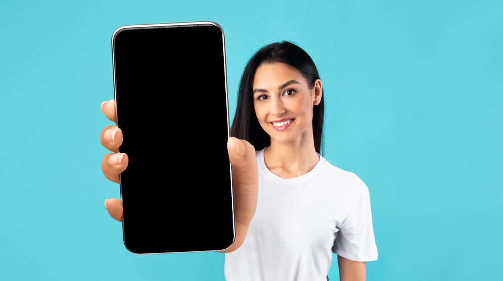 Mobile App Mockup. Beautiful Young Woman Showing Big Smartphone With Black Blank Screen, Smiling Female Demonstrating Free Copy Space For Your Design Or Advertisement, Posing Over Blue Background
