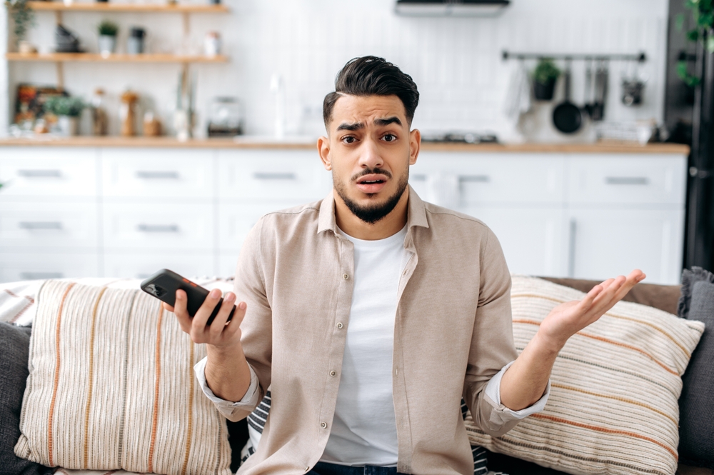 Confused puzzled indian or arabian guy in casual clothes, sits on a sofa in an interior living room, holds a smartphone in his hand, looks questioningly at the camera, spreading his arms around
