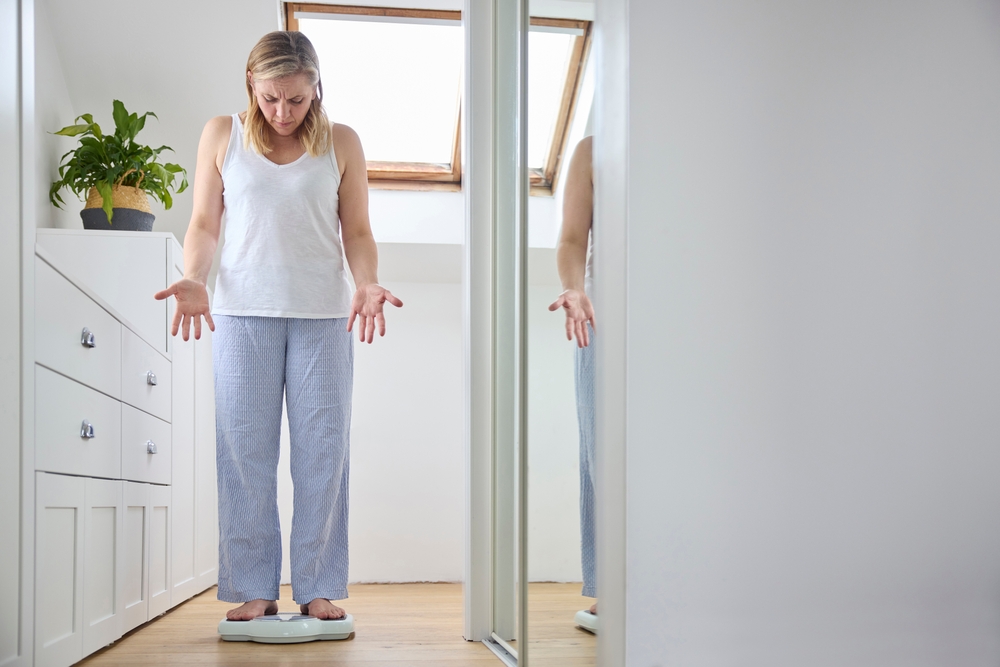 Menopausal Mature Woman Concerned With Weight Gain Standing On Scales In Bedroom At Home
