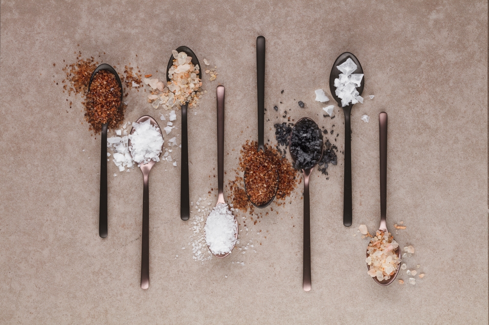 Different kinds of salts in a spoon on a stone table, top view. Composition with different salt on a beige background. Sea salt, cooking salt, Himalayan and black salt .
