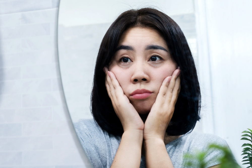 Asian woman looking at a mirror to check skin problems with eyes bags, dark circles, wrinkles and crow's feet on face