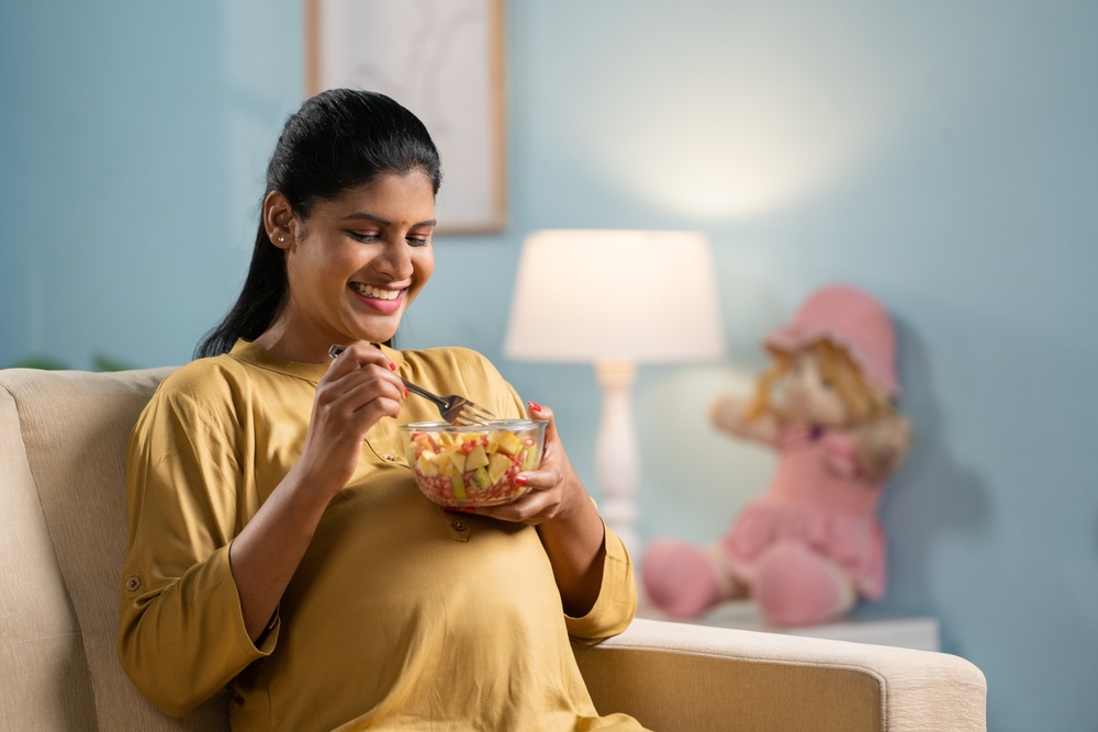 Happy indian pregnant woman eating fruits salad while sitting on sofa at home - concept of healthy lifestyles, Maternal Pregnancy nutrition.
