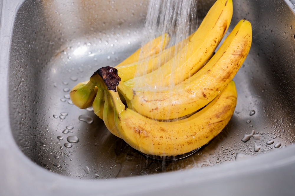 A bunch of ripe bananas being washed under running water in a stainless steel sink, depicting food safety and preparation