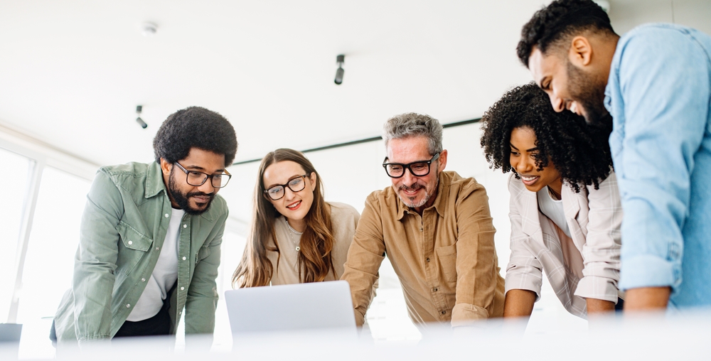 A diverse team of colleagues enthusiastically engages with content on a laptop, capturing a collaborative work culture and an effective brainstorming session in a contemporary office
