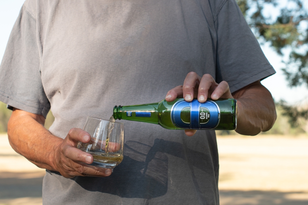 Man pouring a bottle of cold blue label 0.0 alcohol free beer into a glass on a hot sunny day, shadow of bottle and glass on shirt

