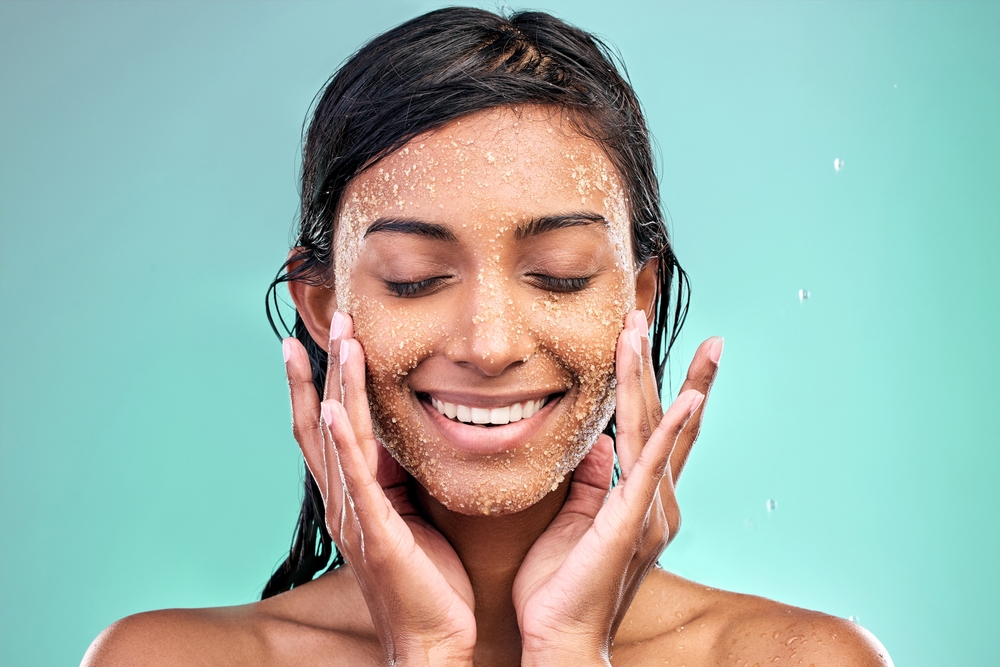 Woman face, beauty and salt scrub in studio with water drops for skin care, hygiene and dermatology. Model eyes closed thinking of facial exfoliation or makeup removal with texture on blue background
