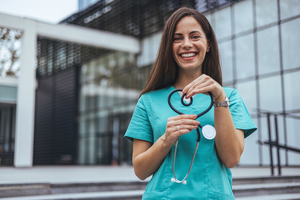 A joyful Caucasian female nurse forms a heart with her stethoscope, displaying care outside a medical facility, dressed in scrubs, symbolizing compassion in healthcare.
