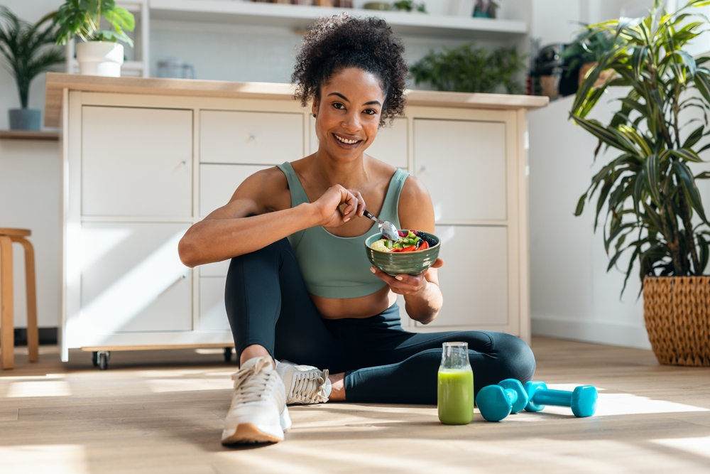 Shot of athletic woman eating a healthy fruit bowl while sitting on floor in the kitchen at home
