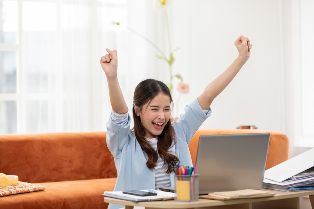 Asian young woman joyfully raises her arms in celebration while working on her laptop at home,moment of professional success, reflecting the positive side of remote work and effective goal accomplish
