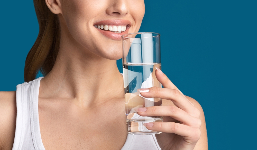 Cropped of woman, dressed in a white tank top, smiling and drinking water from a glass. The background is a solid blue. The image focuses on the womans face, neck, and hand holding the glass