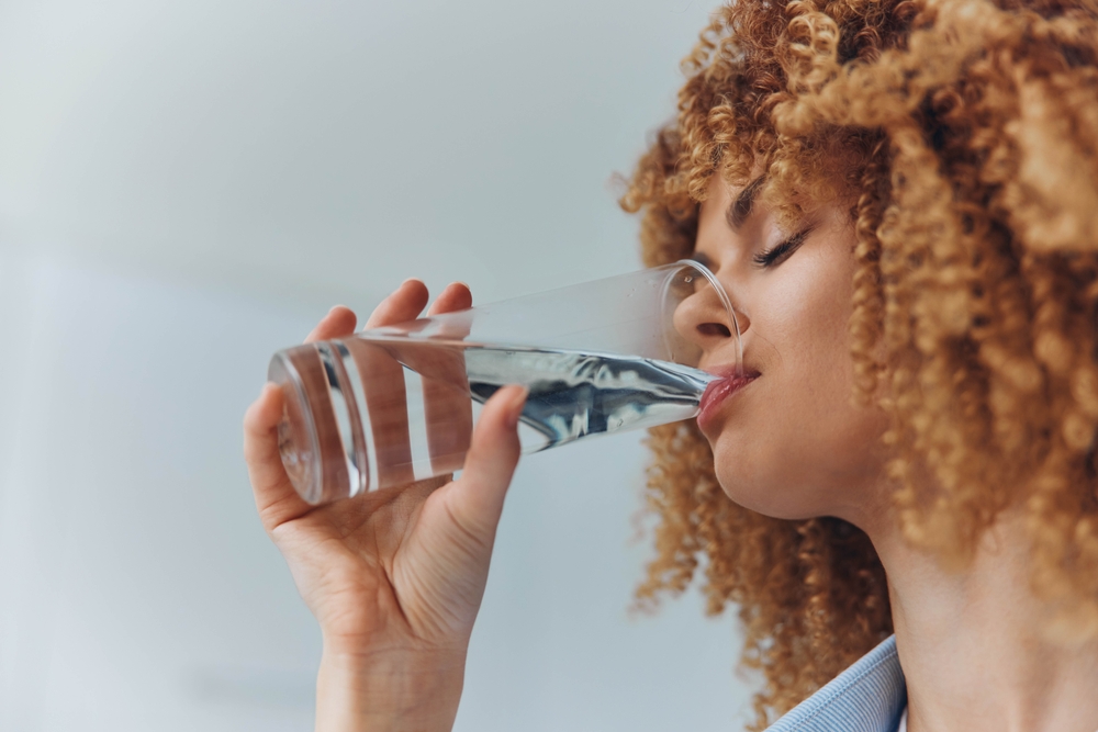 Curly haired woman drinking water from a glass with a refreshing look on her face
