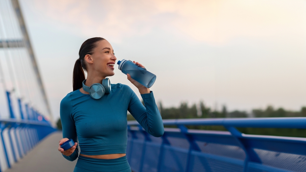 Fit European woman relaxing and drinking water on seaside promenade after running and training outdoors in the evening, panorama, copy space

