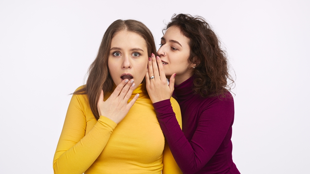 Two women, one in a yellow shirt and the other in a purple shirt, sharing a secret. The woman in yellow shows a surprised expression.