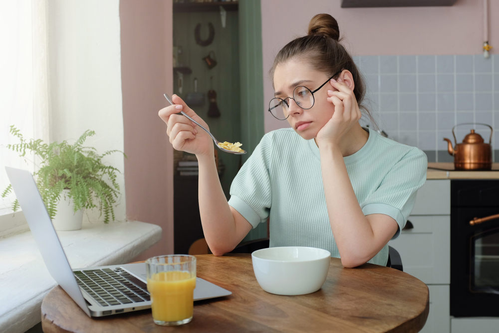 Horizontal portrait of attractive Caucasian girl sitting in kitchen in morning looking tired while eating breakfast, her laptop lying open on table, not sure what to do during day, feeling bored

